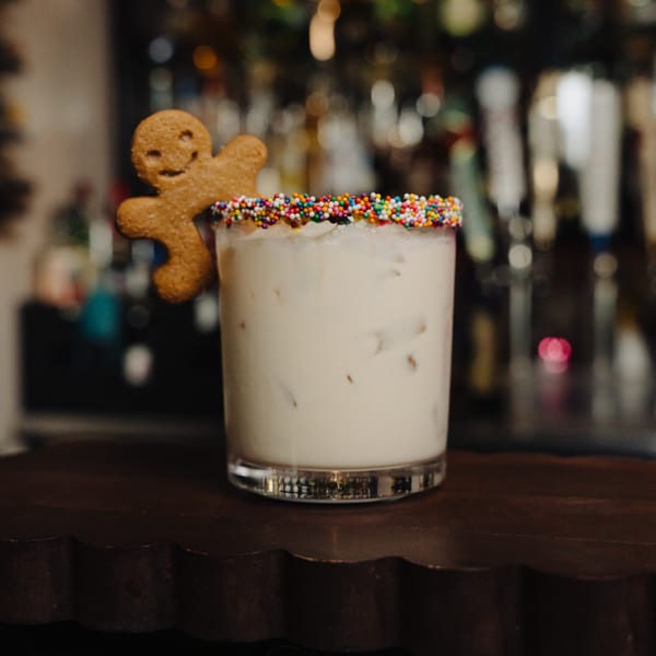 A white cocktail with ice, rainbow sprinkles on the rim, and a gingerbread man cookie garnish.
