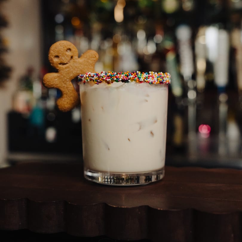 A white cocktail with ice, rainbow sprinkles on the rim, and a gingerbread man cookie garnish.