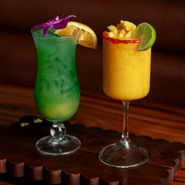 Two vibrant cocktails, one green with an orchid and orange slice, one yellow with mango and lime.