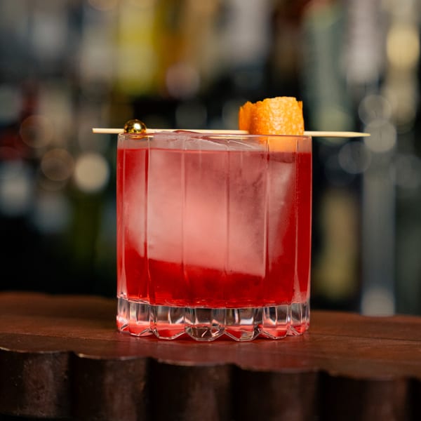 A vibrant red cocktail with a large ice cube, garnished with an orange peel and gold sphere.