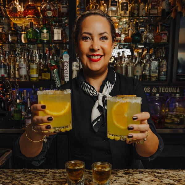 Smiling bartender holding two margaritas with orange slices and salt rims, with liquor bottles in background.