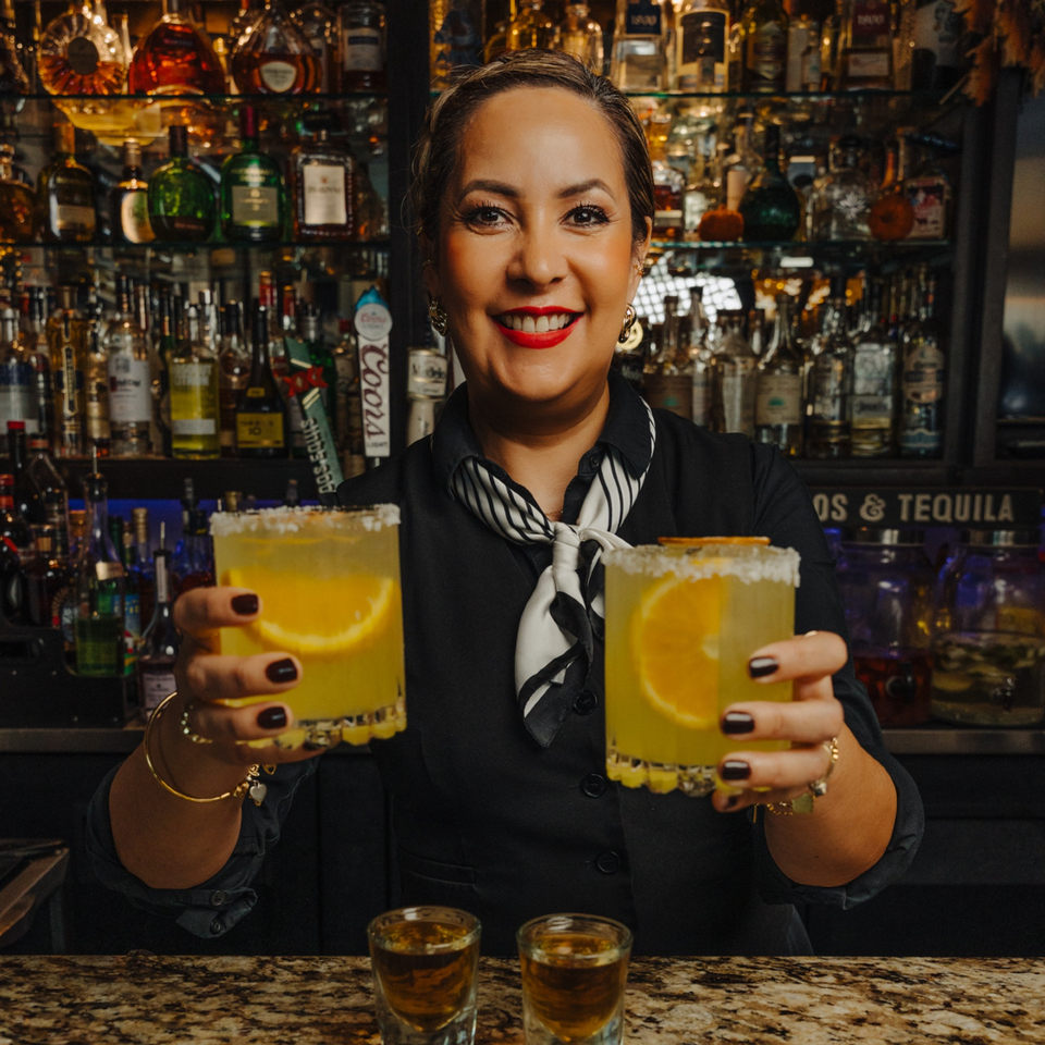 Smiling bartender holding two margaritas with orange slices and salt rims, with liquor bottles in background.