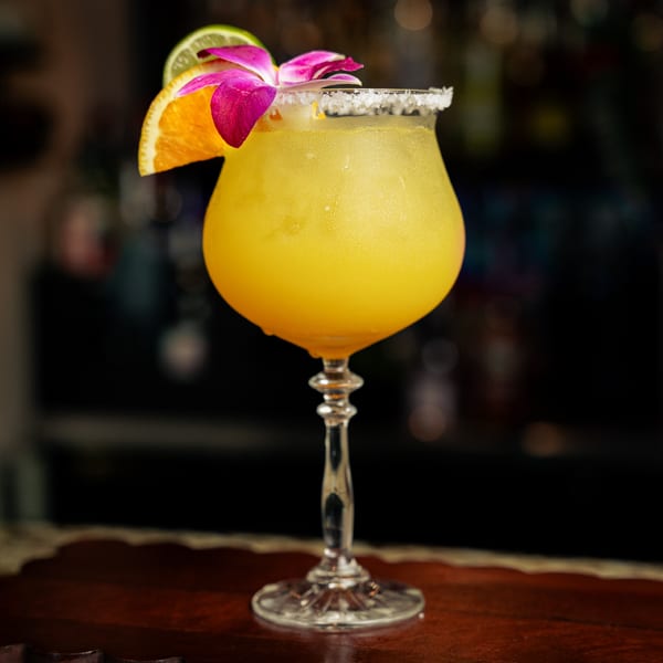 A vibrant yellow El Tarasco margarita, garnished with an orange slice, lime, and purple orchid, on a bar.