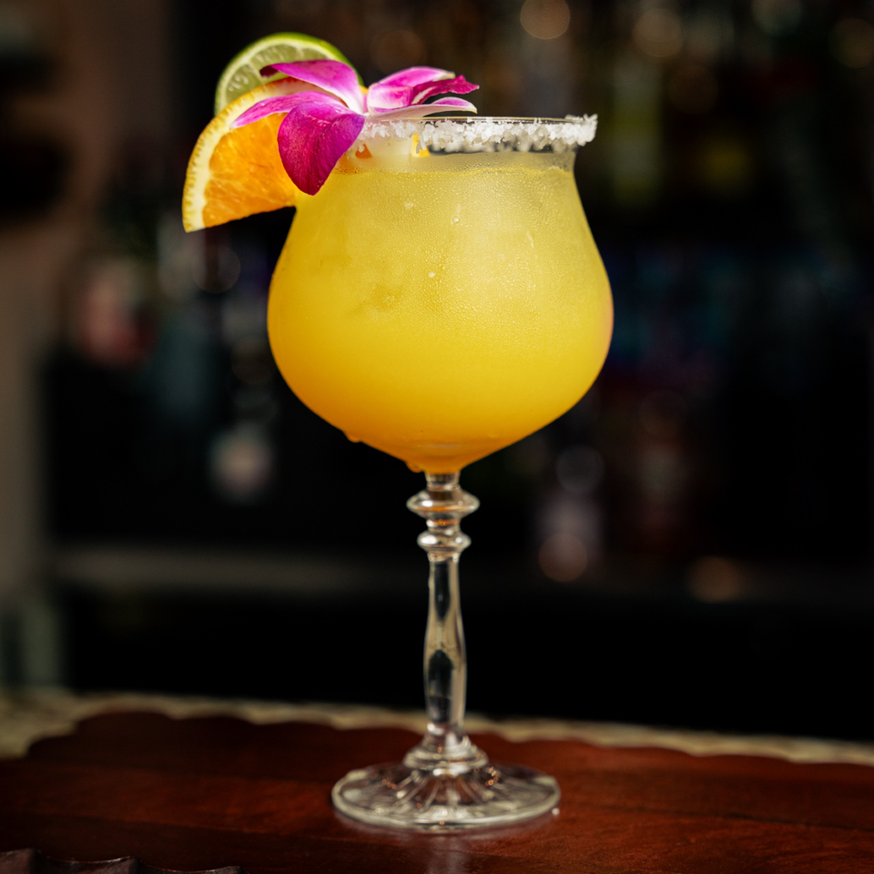 A vibrant yellow El Tarasco margarita, garnished with an orange slice, lime, and purple orchid, on a bar.
