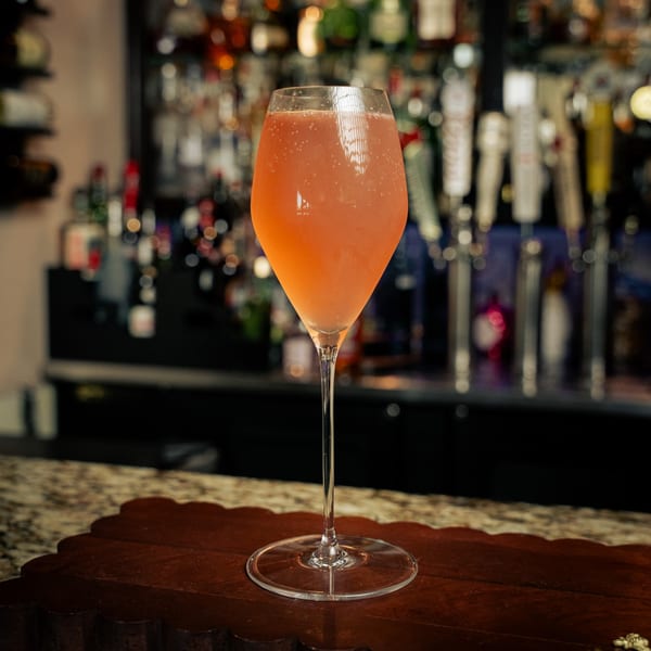 A tall, elegant glass of sparkling pink drink sits on a wooden tray at a bar.