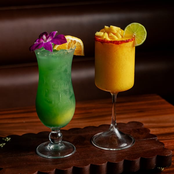 Two vibrant Mexican cocktails: a green drink with an orchid and orange slice, and a mango drink with chili and lime.