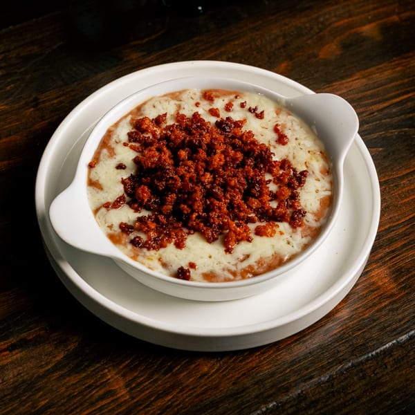 A white bowl of melted cheese and chorizo, served on a white plate on a dark wooden table.