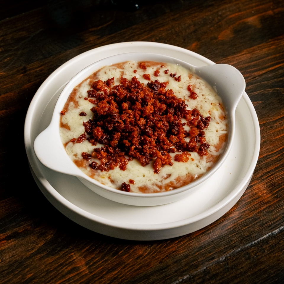 A white bowl of melted cheese and chorizo, served on a white plate on a dark wooden table.