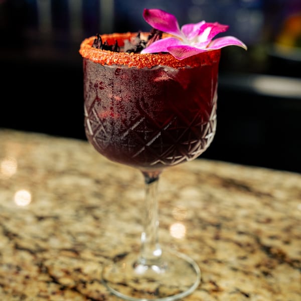 A vibrant red El Tarasco cocktail in a patterned glass, garnished with a pink orchid and chili salt rim.