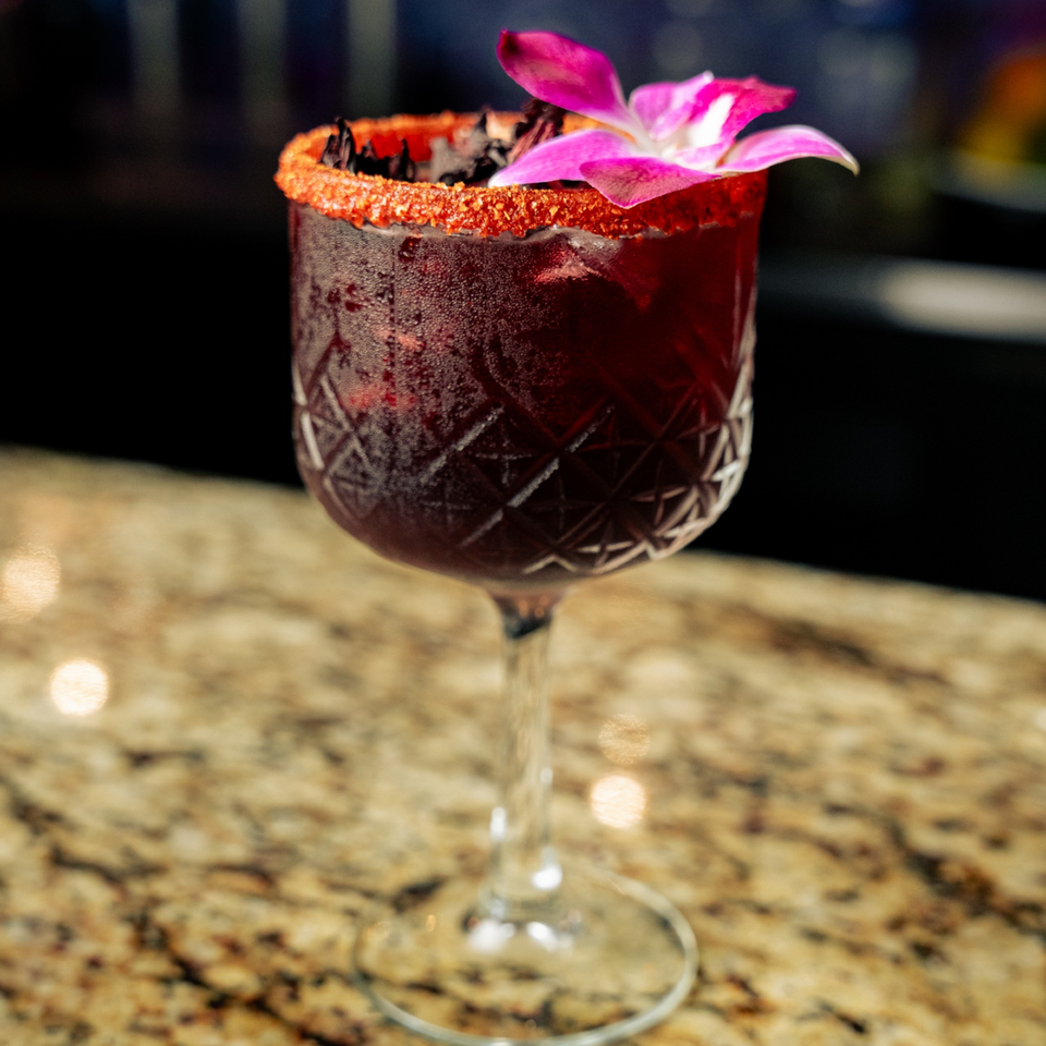 A vibrant red El Tarasco cocktail in a patterned glass, garnished with a pink orchid and chili salt rim.