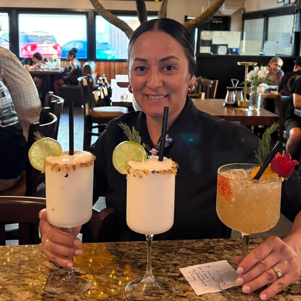 A smiling woman holds two El Tarasco Restaurant Mooney margaritas and a cocktail at a bar.