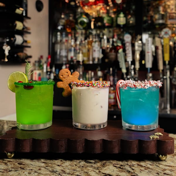 Three festive holiday cocktails, green, white, and blue, on a bar with bottles in the background.