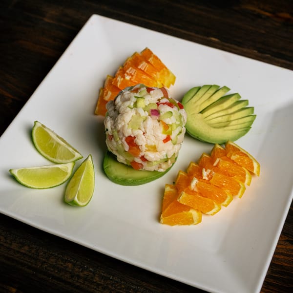 Ceviche with diced vegetables, surrounded by avocado slices, orange segments, and lime wedges on a white plate.