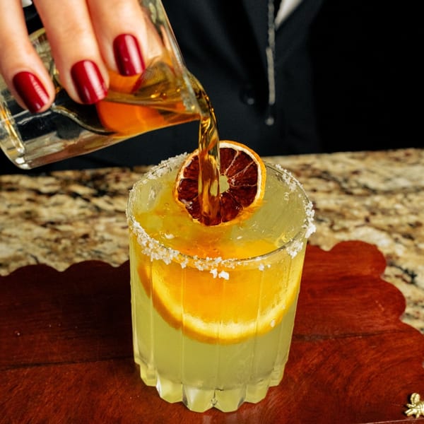 A hand with red nails pours amber liquid into a margarita with orange slices and a salted rim.